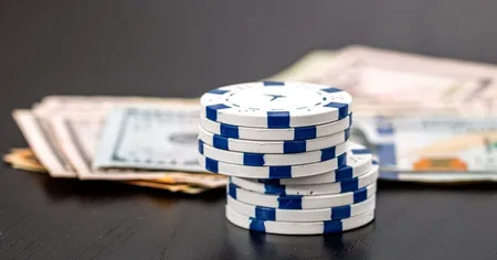 Stacks of blue and white poker chips are placed in front of a pile of U.S. dollar bills on a dark wooden surface.