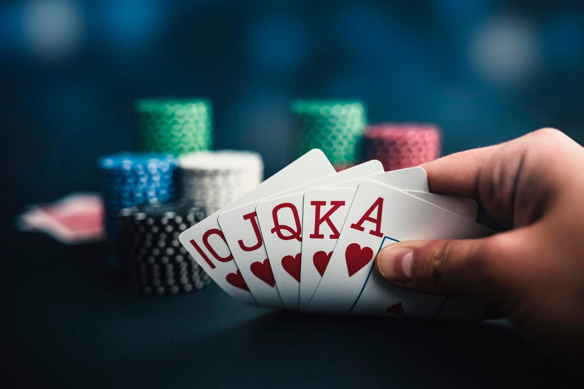 Hand holding a royal flush in hearts, with poker chips stacked in the background on a blue table.