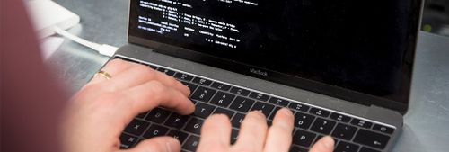 Närbild på två händer som skriver på en laptop, på skärmen syns vit text på svart bakgrund, texten är kod