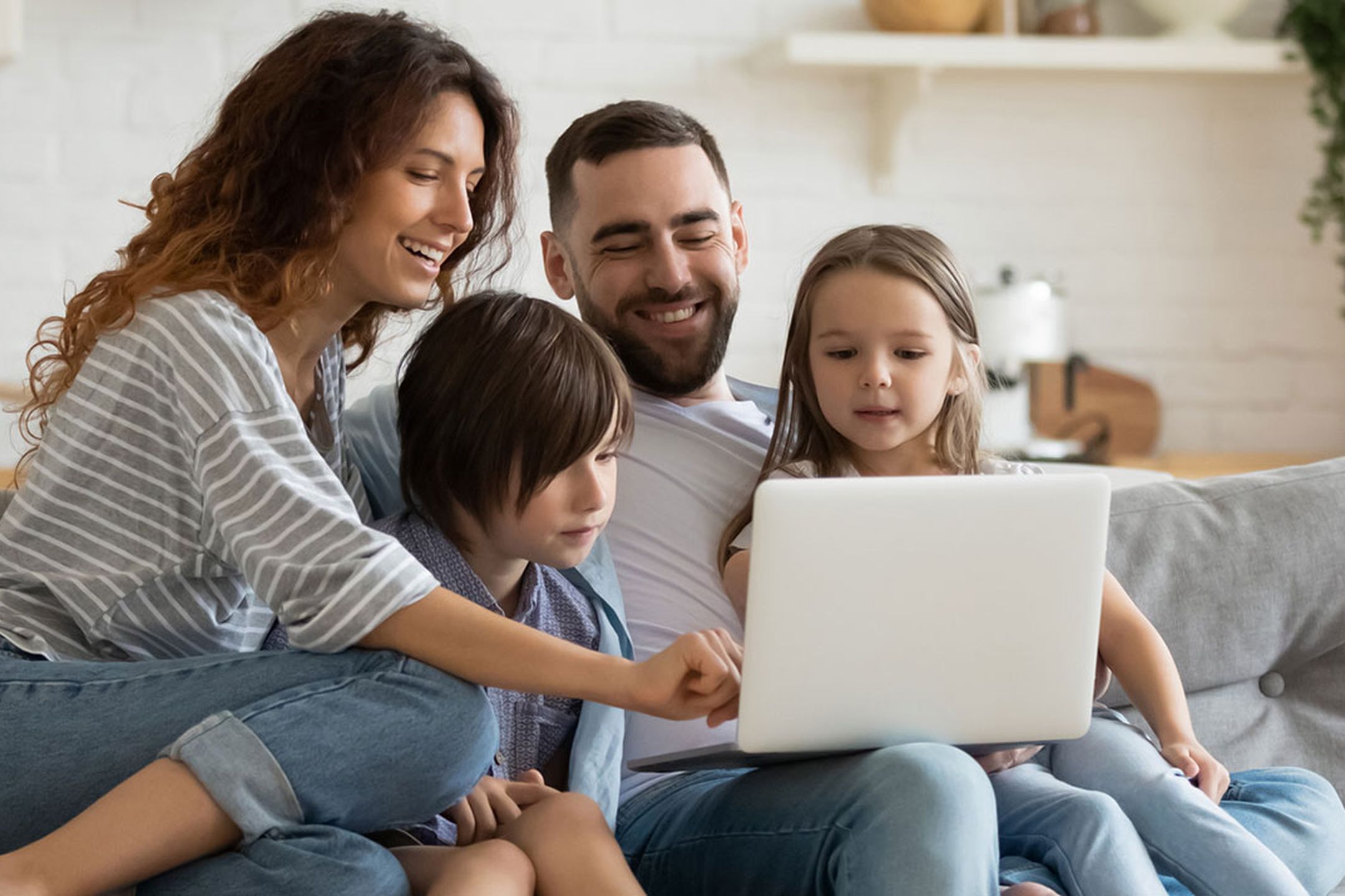 En familj bestående av en två vuxna personer och två små barn som sitter i en soffa. De sitter tätt ihop och tittar på en laptop. De vuxna ler och ser glada ut.