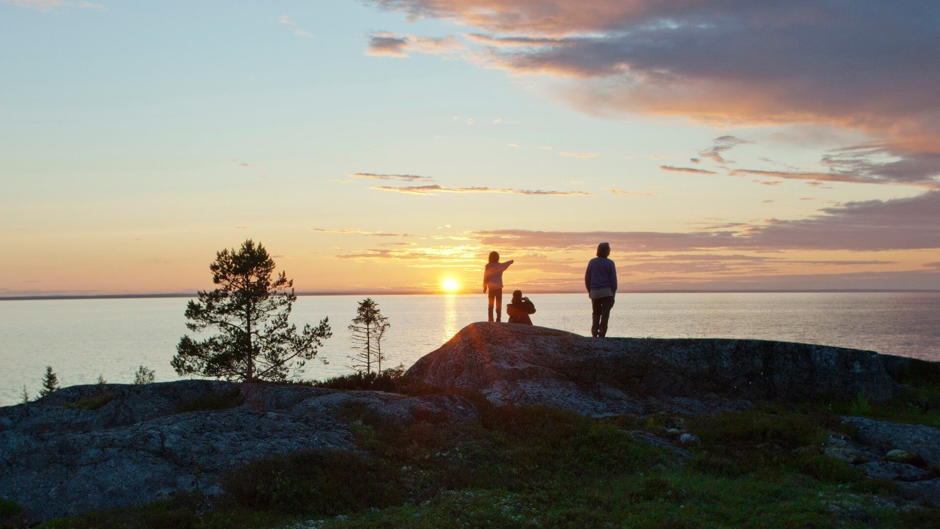 Tre personer som står på en klippa vid havet. I bakgrunden syns havshorisonten där solen håller på att gå ner.