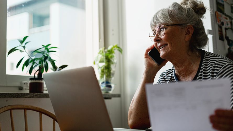 Kvinna sitter vid skrivbord med växter dator och dokument medan hon pratar i telefon