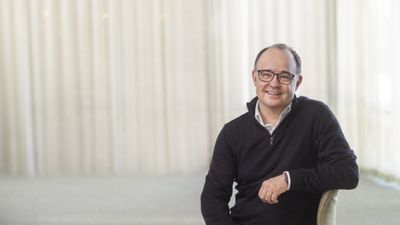 Leende man med glasögon och svart tröja sitter inomhus framför ljust draperi