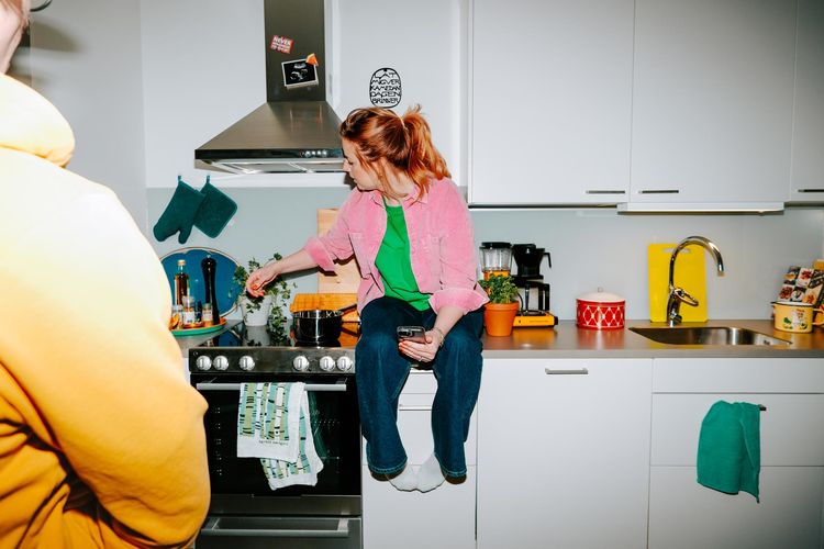 En person i rosa skjorta och jeans sitter på köksbänken och kryddar en gryta samtidigt som hen håller en mobil