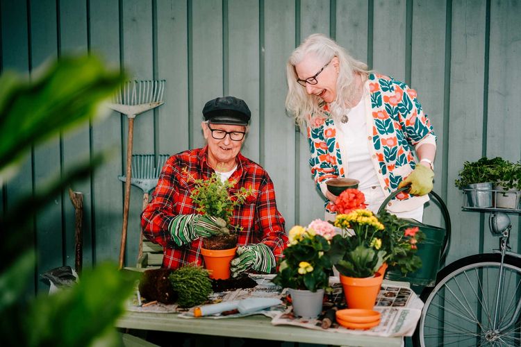 Två äldre personer ler och planterar färgglada blommor vid ett trädgårdsbord, omgivna av verktyg och växter