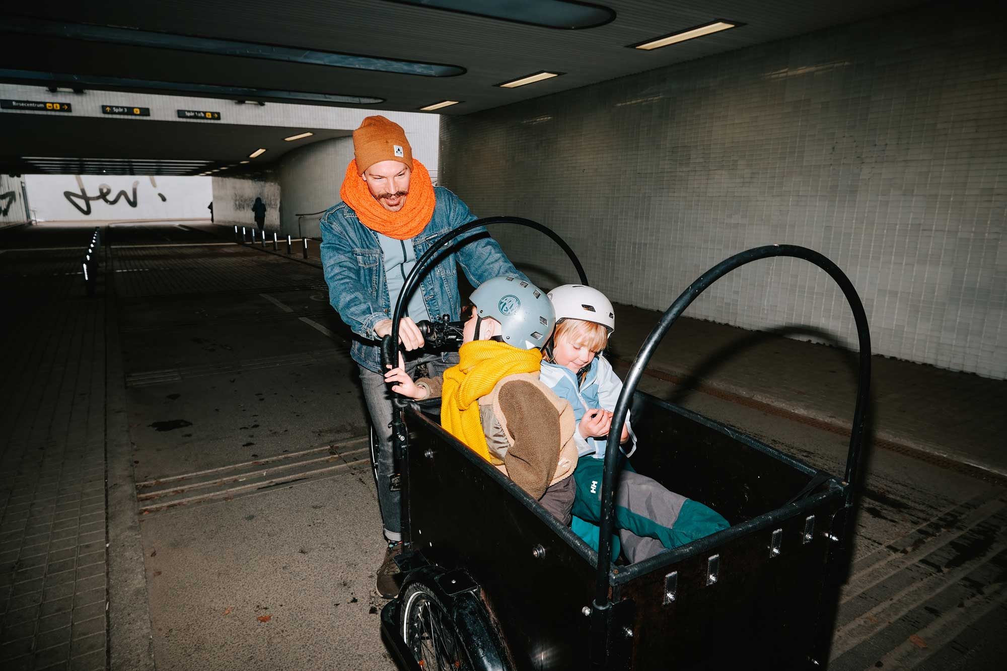 En vuxen med orange halsduk cyklar med två barn i en lådcykel genom en tunnel i Umeå