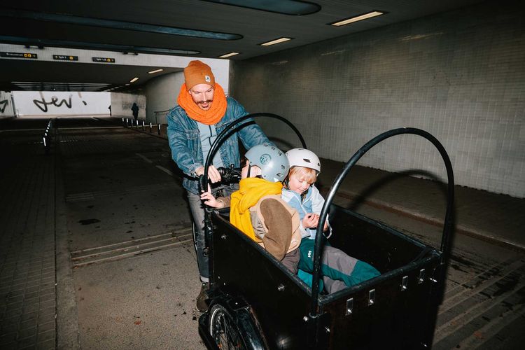 En vuxen med orange halsduk cyklar med två barn i en lådcykel genom en tunnel i Umeå