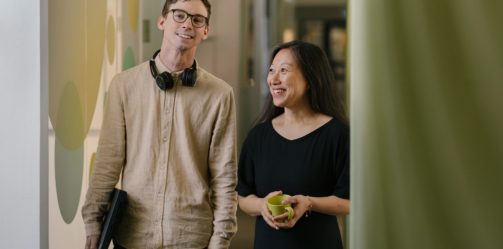 Två personer som står i en företagskorridor. Den ena personen håller i en kaffekopp, den andra personen har ett par hörlurar runt halsen och håller i en laptop i ena handen. Personerna ler.