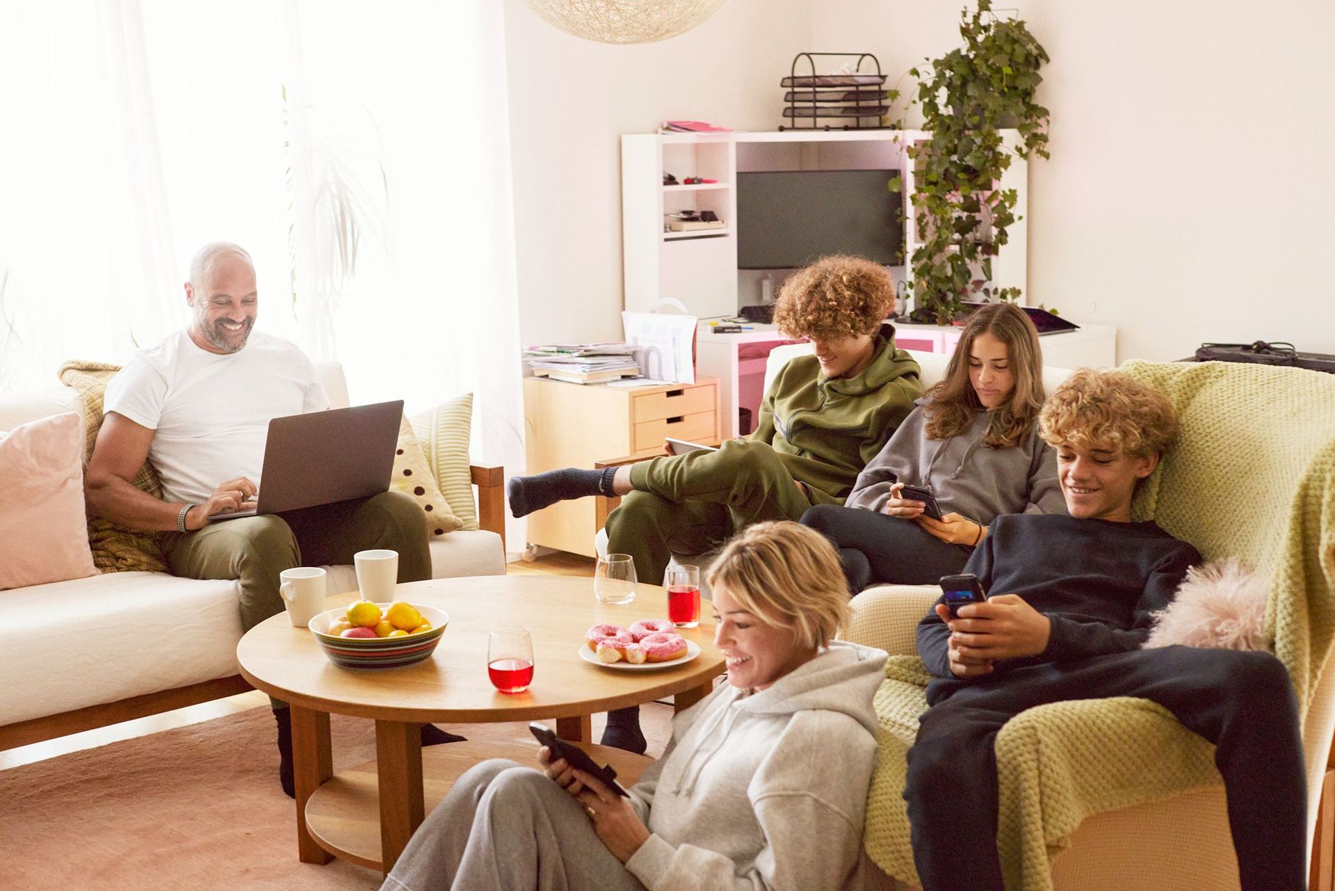 Familj som sitter med olika digitala enheter i soffa, fåtöljer och på golvet