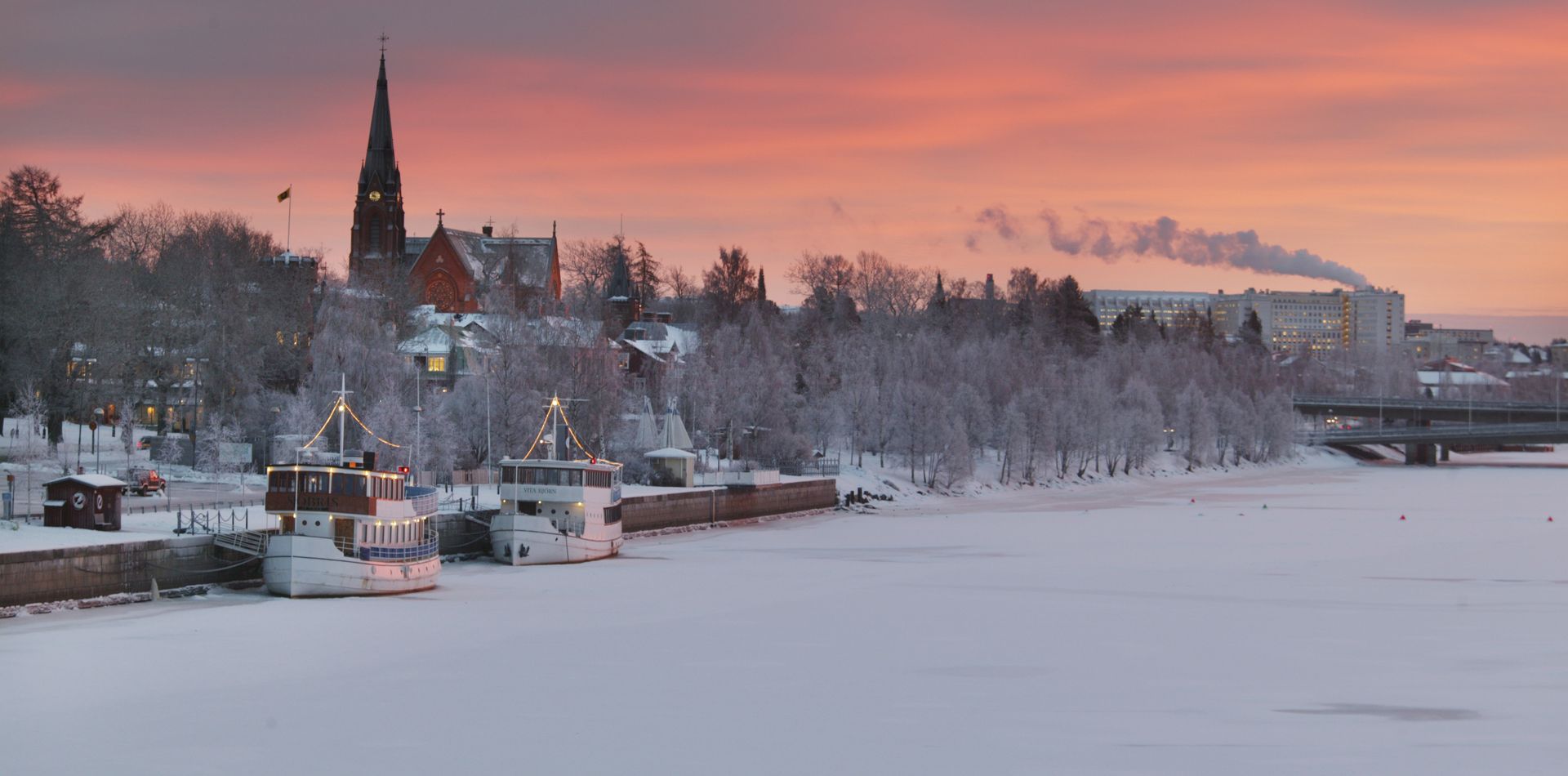 Vy över Umeå älv vintertid och strandkant med båtar, hus och kyrka, rosa himmel