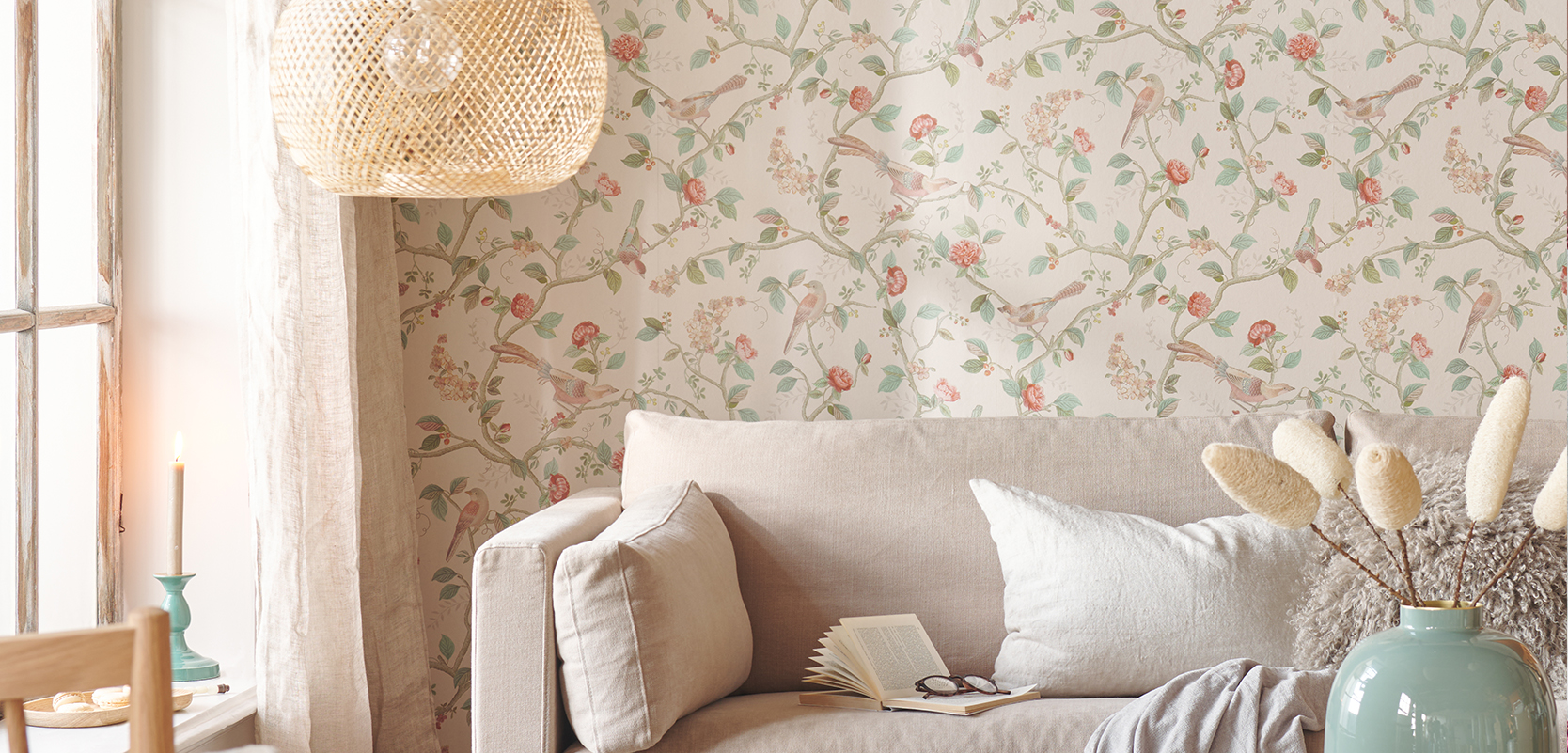 Cozy living room with a beige sofa, floral wallpaper, an open book, and a rattan pendant light. A vase of dried flowers sits nearby.