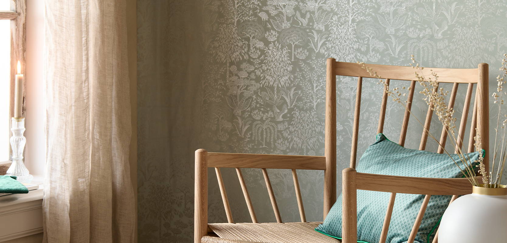 Wooden chair with green cushions beside a window with sheer curtains. Delicate floral wallpaper in the background and a white vase on a table.