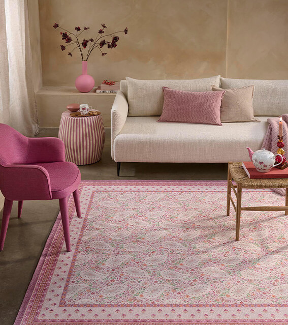 Cozy living room with pink accents featuring a beige sofa, pink armchair, patterned rug, and a wooden coffee table with a teapot set.