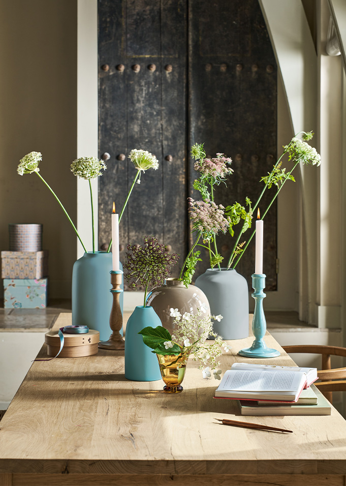 A wooden table with blue vases holding white flowers, candles, books, and decorative boxes against a rustic door backdrop.