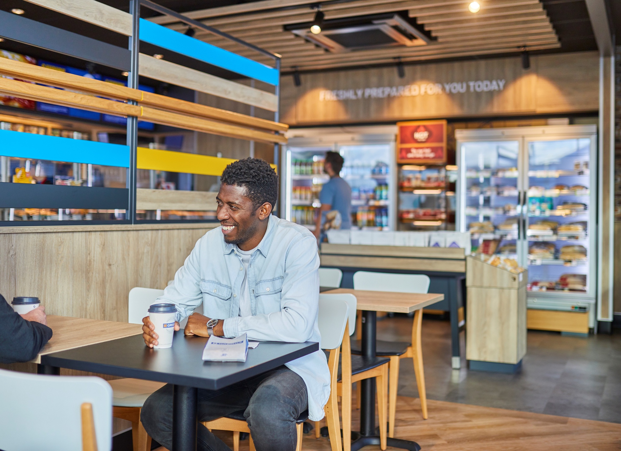 A man sits smiling drinking a coffee in a Greggs store