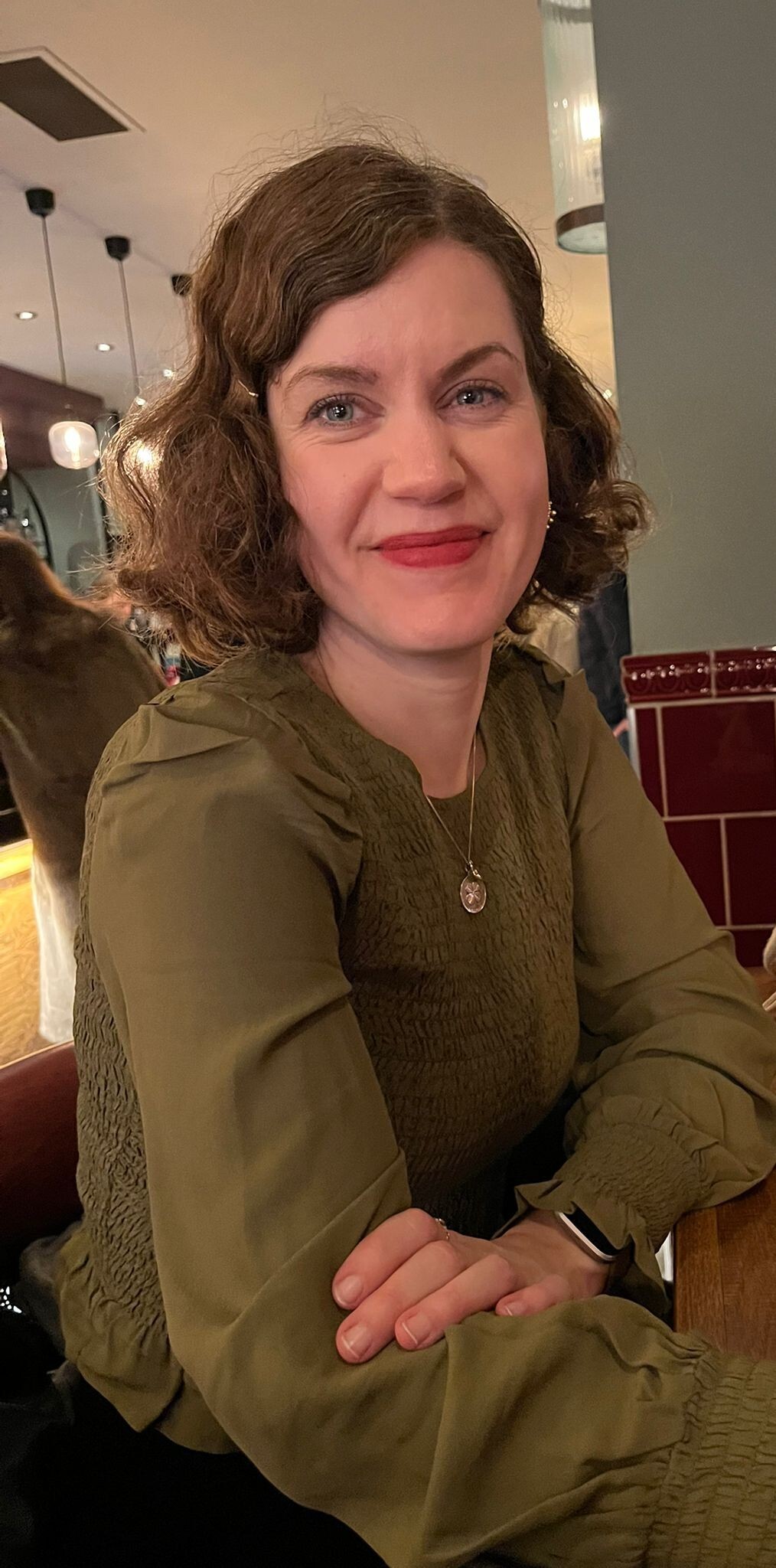 Woman with curly hair and green blouse smiling while seated in a warmly lit restaurant.