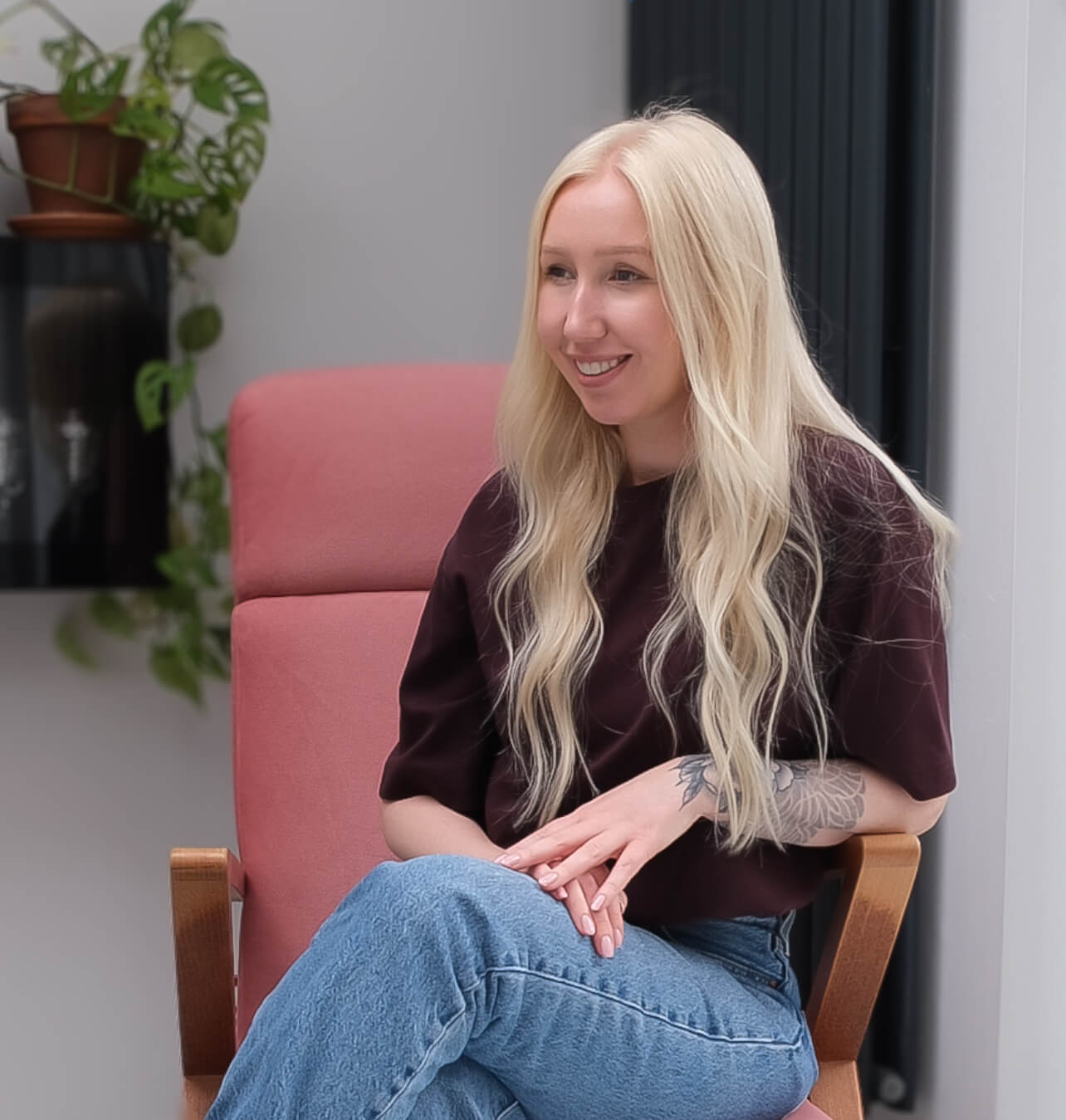 Smiling woman with long blonde hair sits on a pink chair, wearing a dark shirt and jeans.
