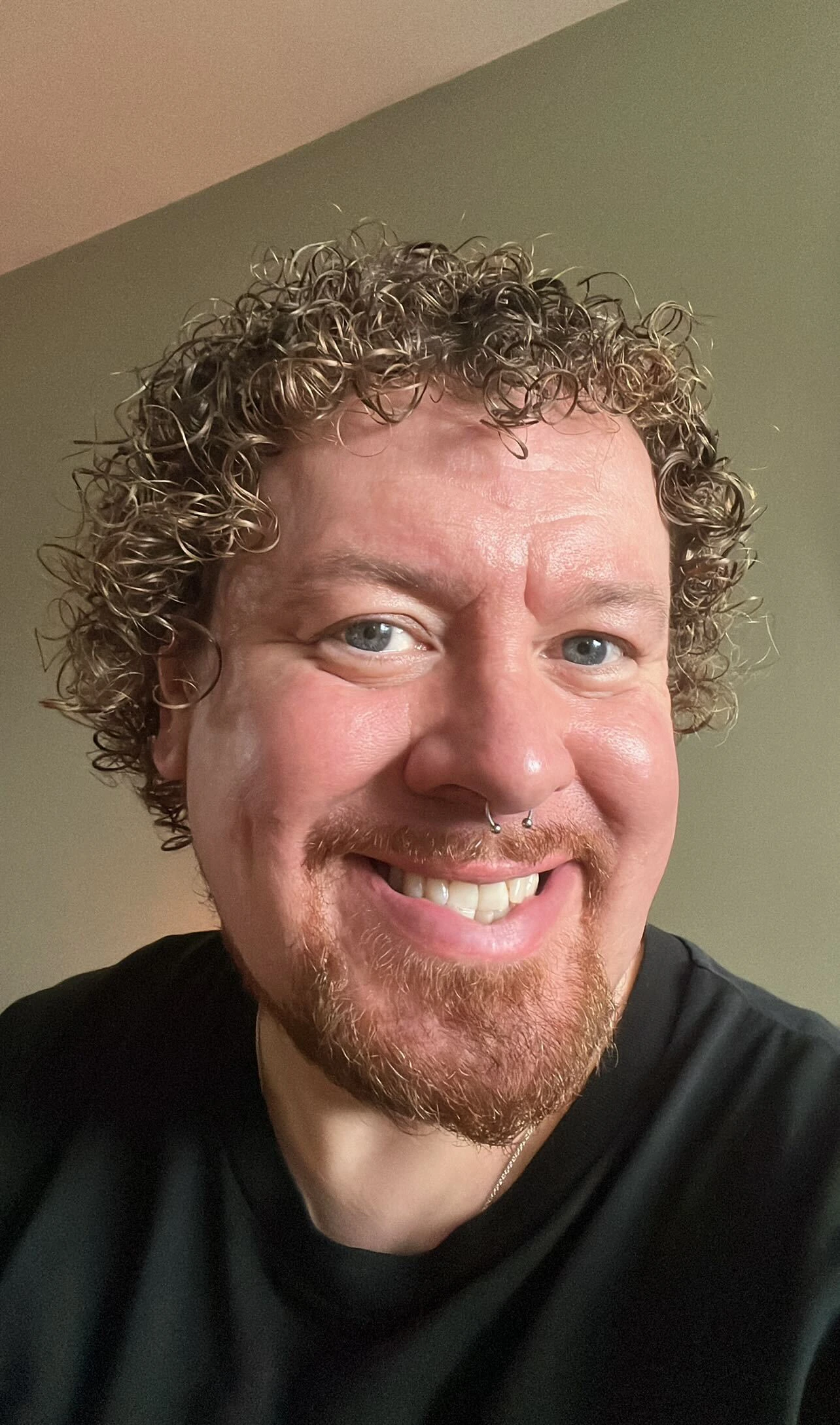Person with curly hair and a beard smiling, wearing a black shirt, against a plain background.