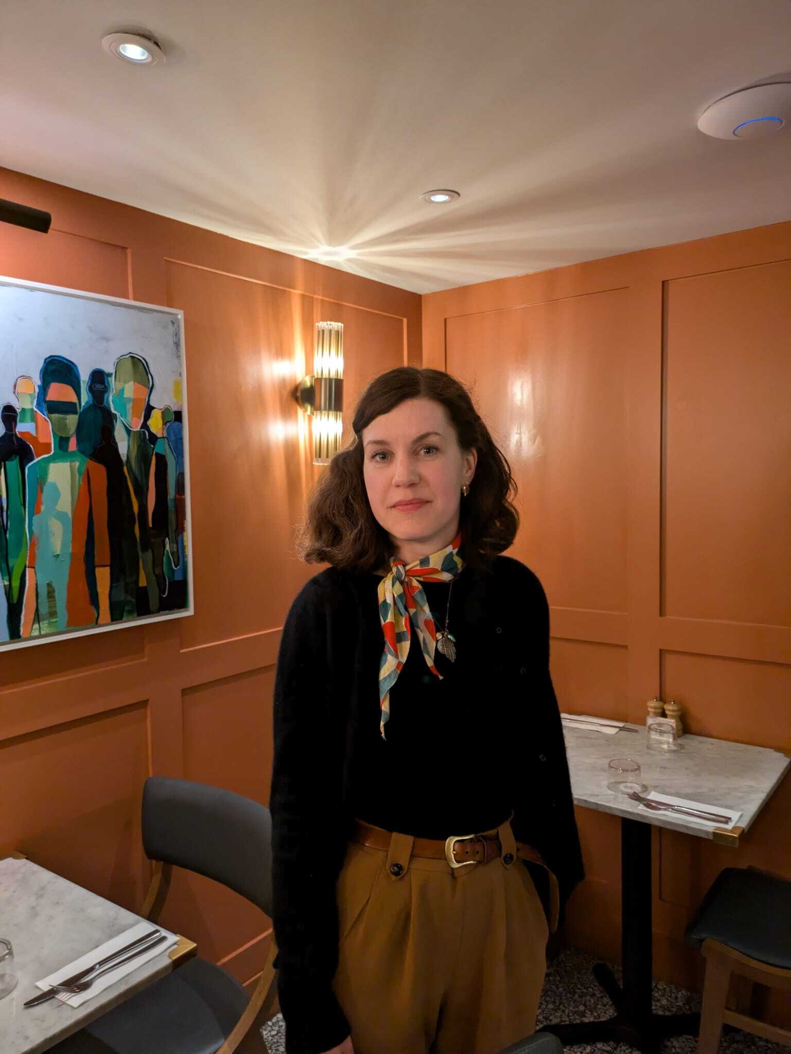 Woman in a black sweater and colourful scarf stands in a warmly lit restaurant.
