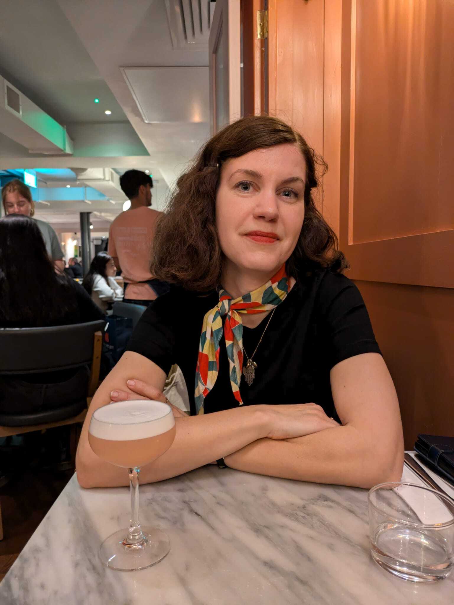 Woman with curly hair and a colourful scarf sits at a marble table with a cocktail.