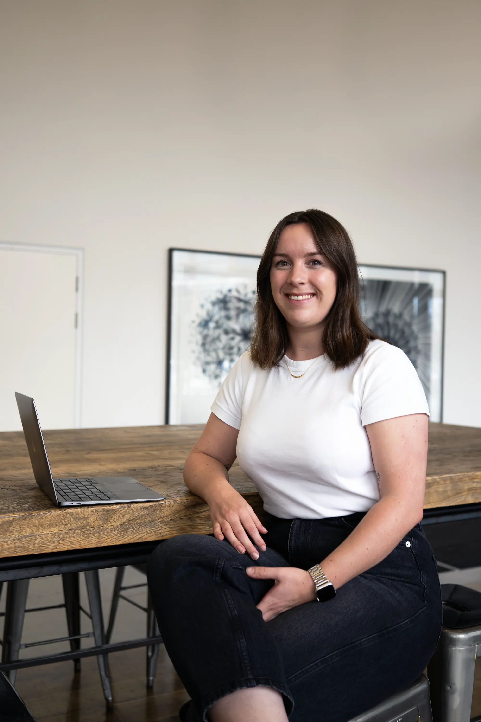 A woman in a white top and black trousers sits at a wooden table with a laptop, smiling.