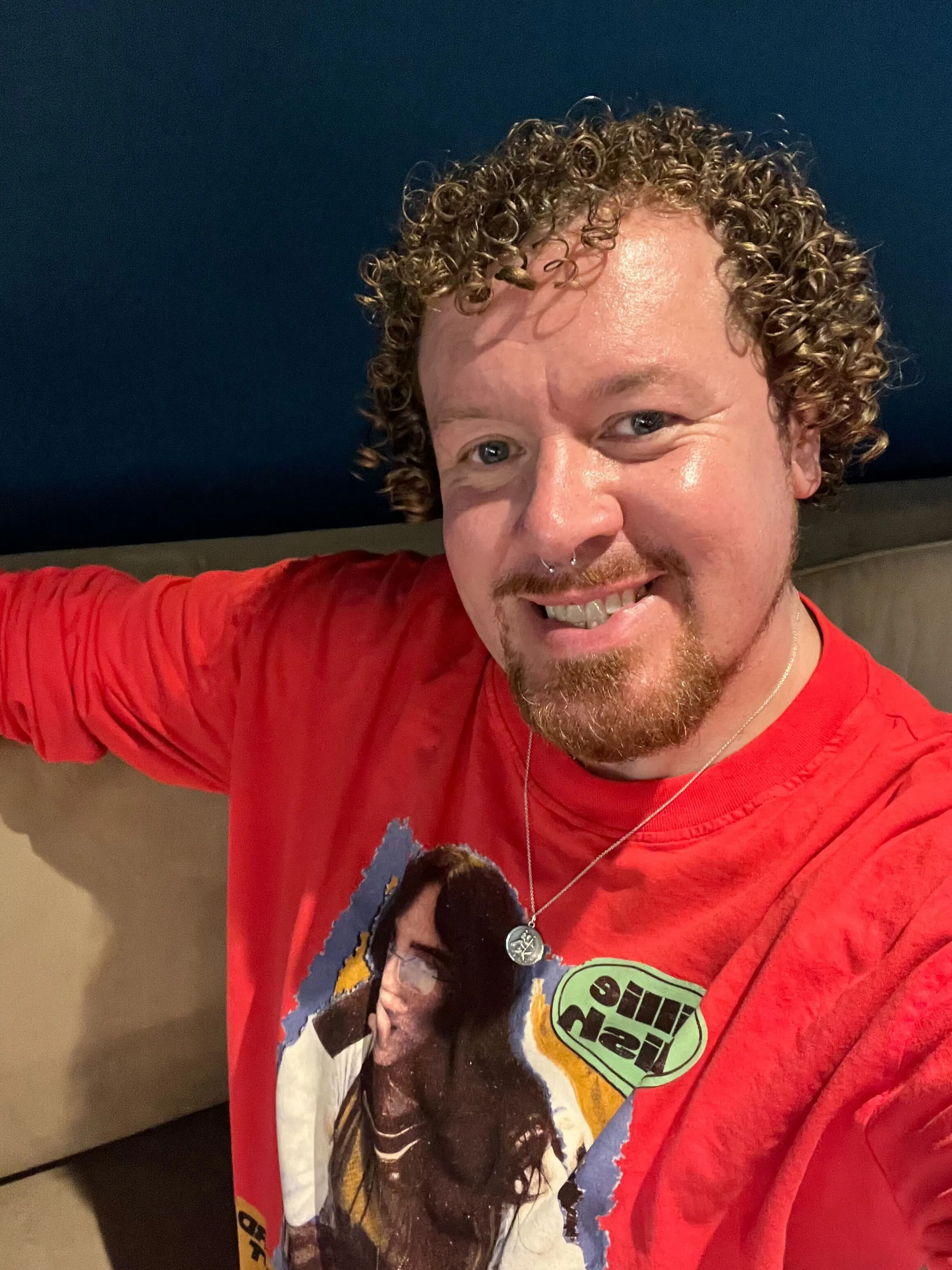 Man with curly hair and beard smiling, wearing a red Billie Eilish shirt and a necklace.