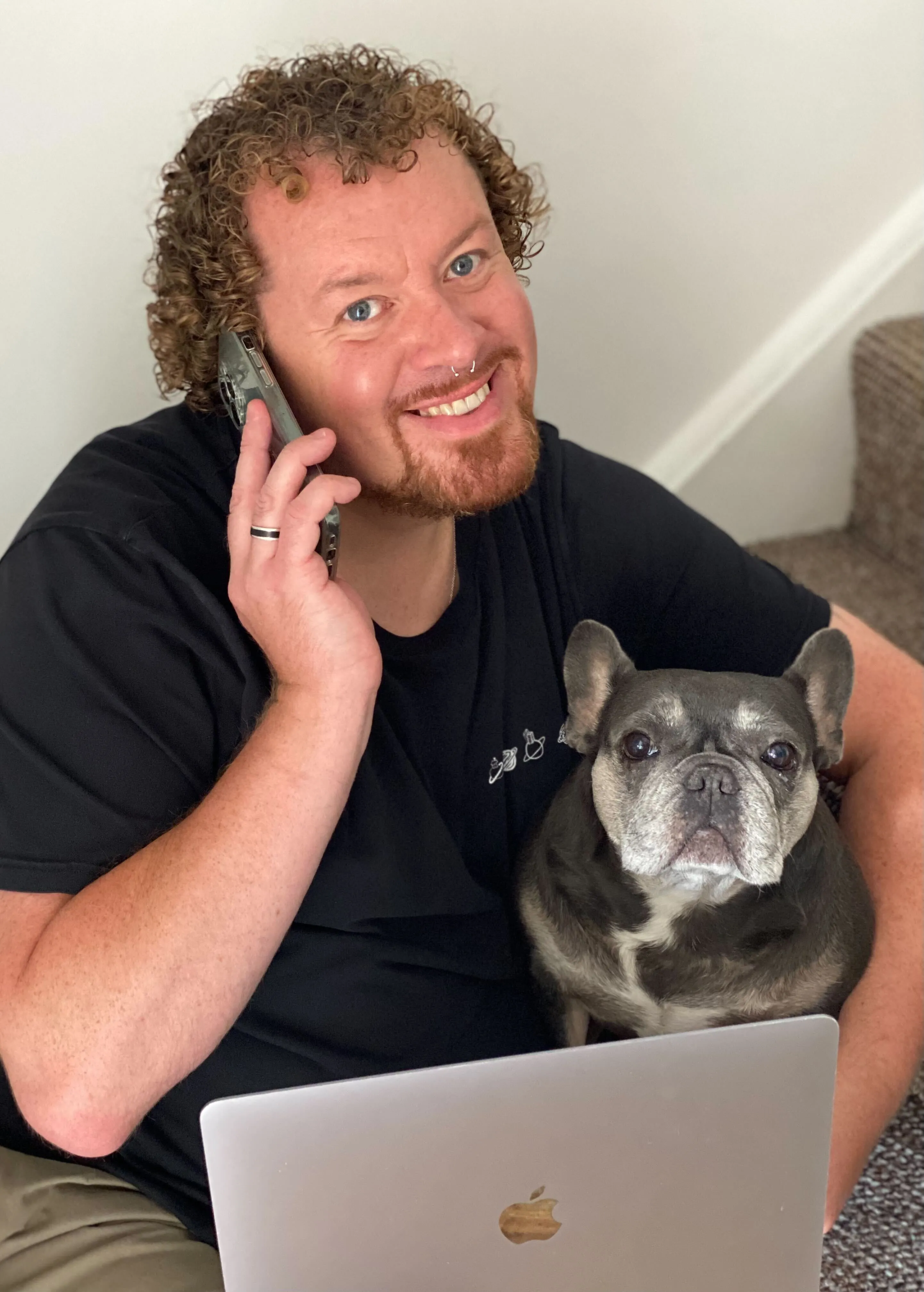Man with curly hair smiling, talking on phone, sitting on stairs with a laptop and a French Bulldog on his lap.