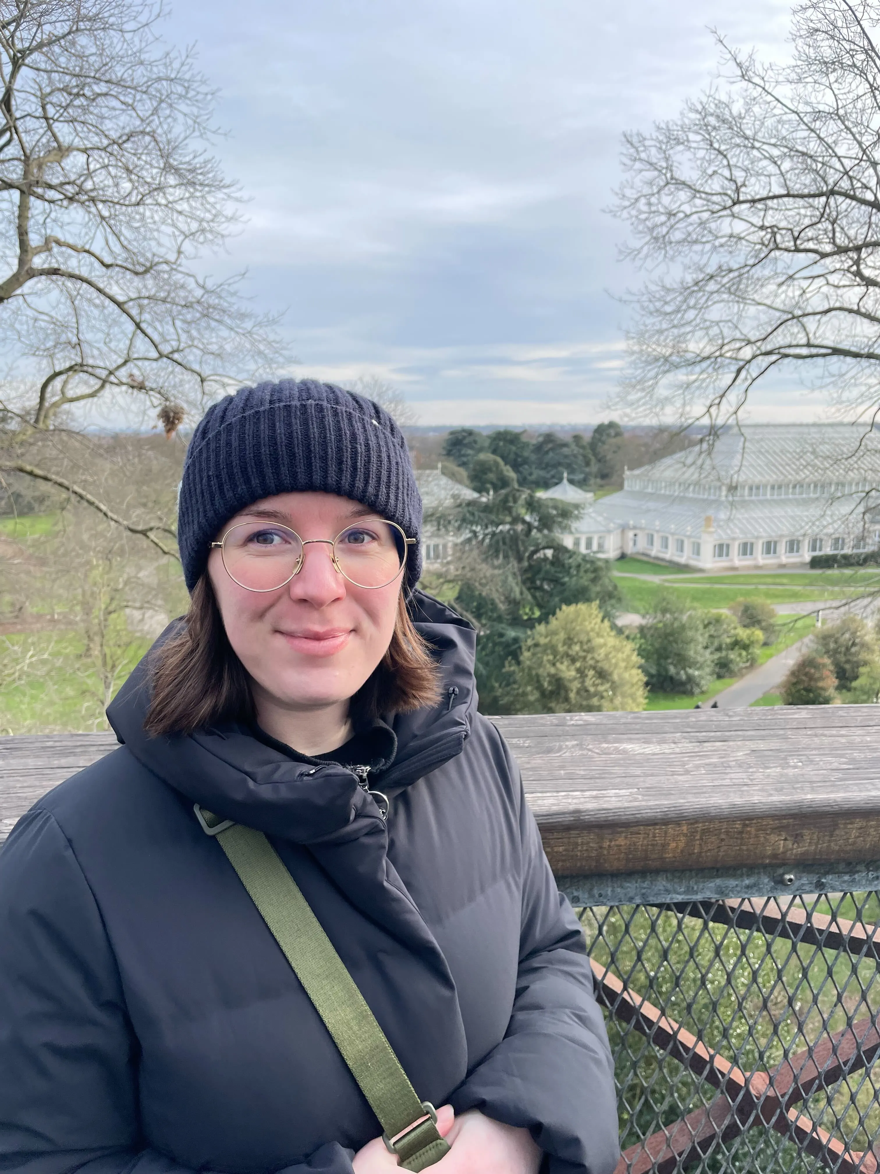 Woman in a black jacket and beanie stands on a wooden platform with a large greenhouse and trees in the background.