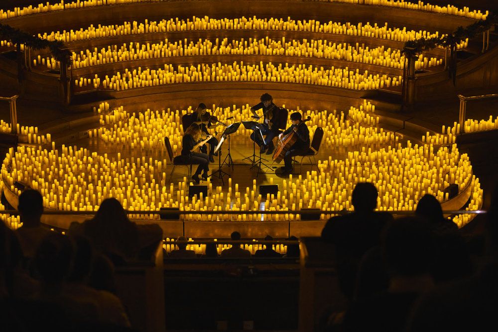 A string quartet performs on a circular stage surrounded by numerous lit candles, with an audience watching in a dimly lit venue.