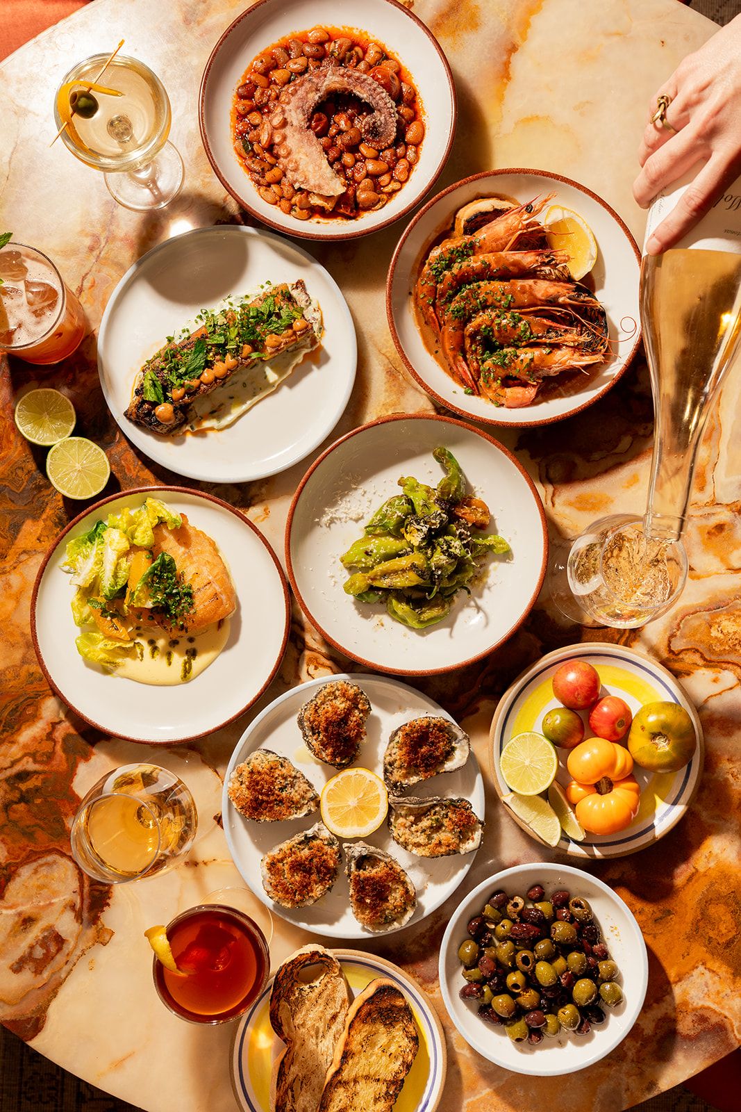 A table with various dishes: seafood, grilled vegetables, oysters, salad, and drinks. A hand pours champagne into a glass.