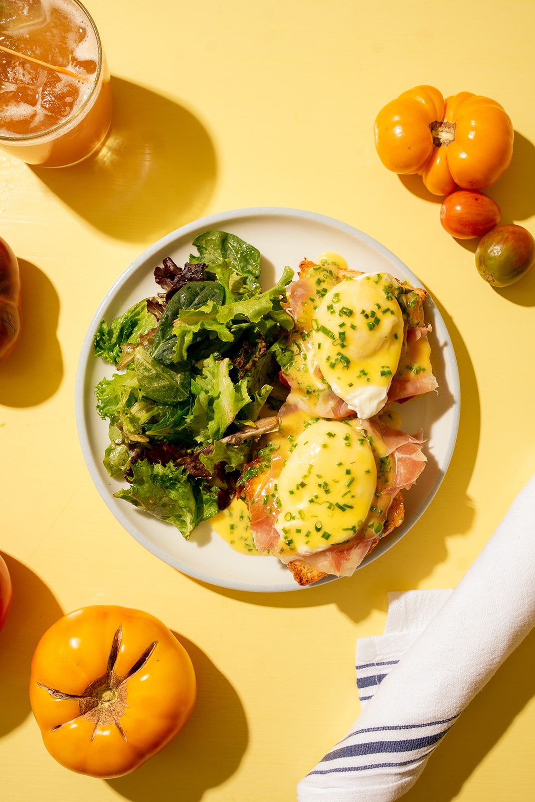 Plate of eggs Benedict with ham and hollandaise, served with mixed greens. Yellow tomatoes and a drink are beside the plate on a yellow table.