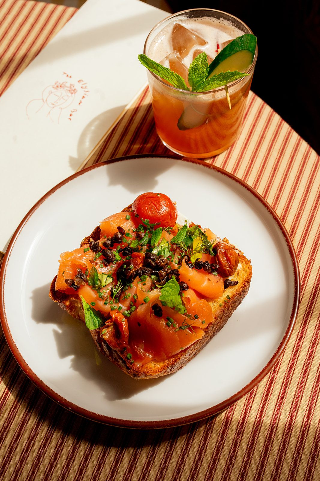 A plate with smoked salmon toast garnished with herbs and tomatoes, next to a cocktail with mint leaves, on a striped tablecloth.