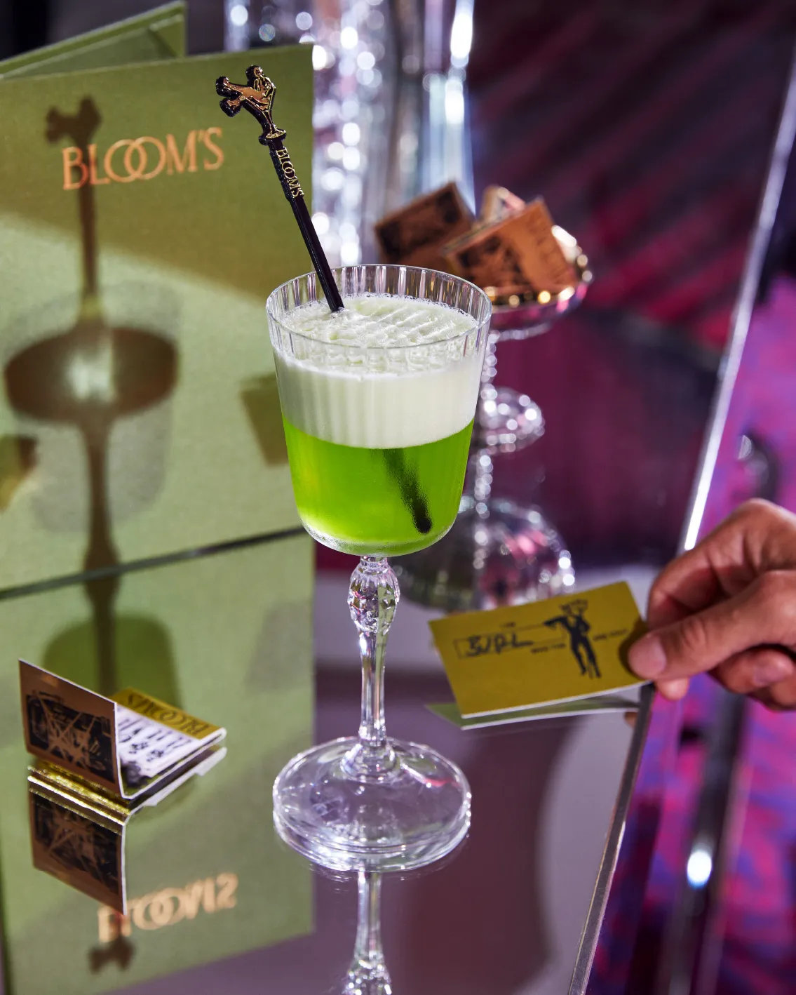 Elegant green cocktail in a crystal glass with a decorative stirrer, set on a reflective surface with a hand holding a card nearby.