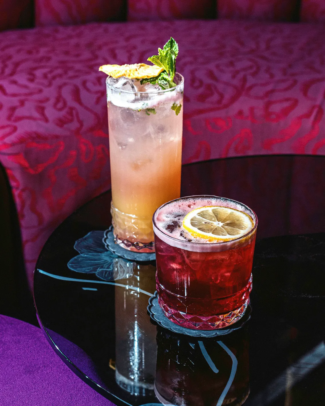 Two cocktails on a dark table: a tall pink drink with mint and a short red drink with a lemon slice, set against a plush red background.
