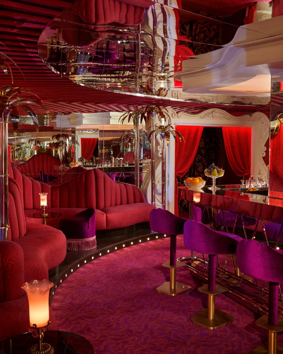 Luxurious bar with red and purple decor, plush seating, mirrored ceiling, and ornate lighting, creating an opulent, retro ambiance.
