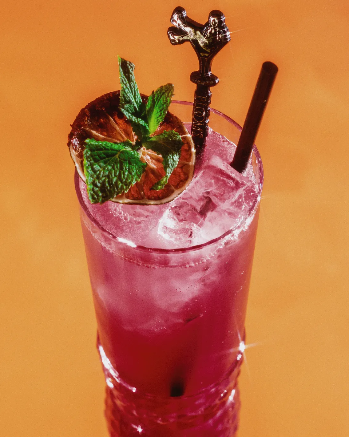 A vibrant pink cocktail with ice, garnished with a mint sprig and dried lime slice, served with a straw and decorative stirrer on an orange background.