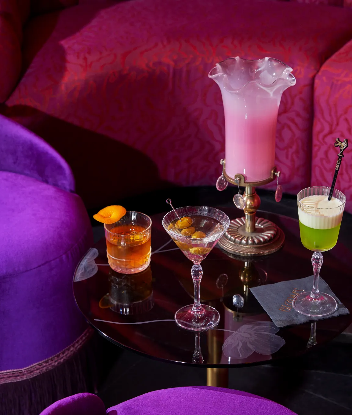 A stylish table with three cocktails and a pink vase, set against vibrant purple and pink seating in a chic, dimly lit lounge.