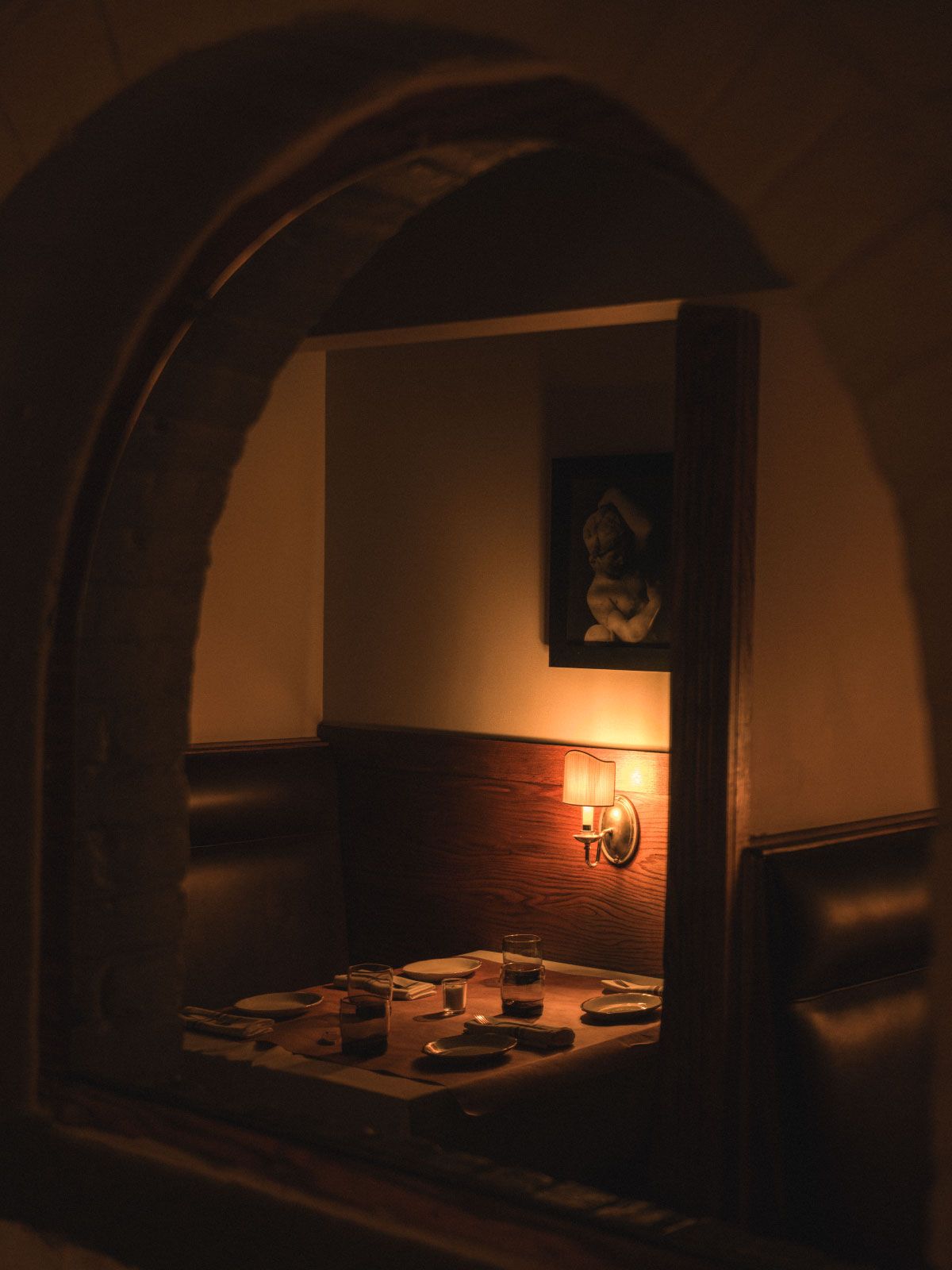 Cozy dimly lit restaurant booth with a wooden table, set with glasses and plates, beneath a soft wall light and framed art.
