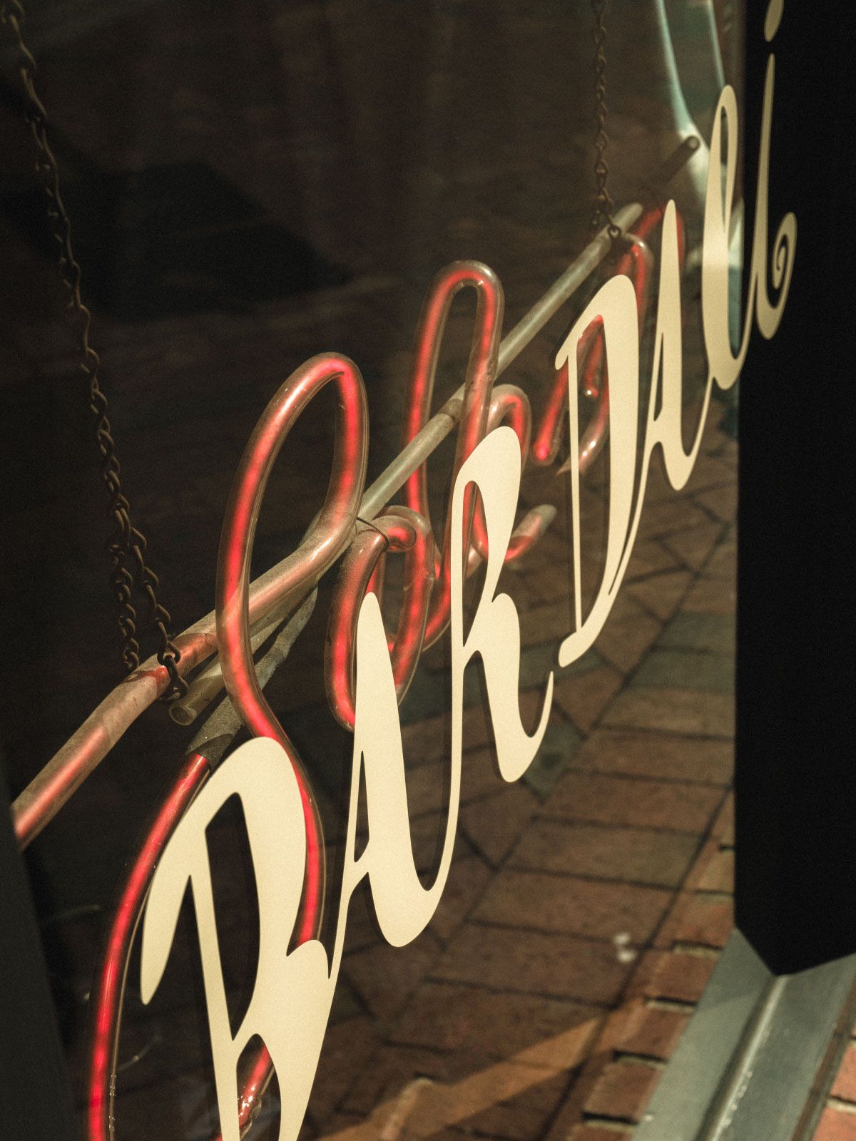 Vintage-style sign reading "Bar Deli" hanging in a window, with red neon light reflecting on the glass.