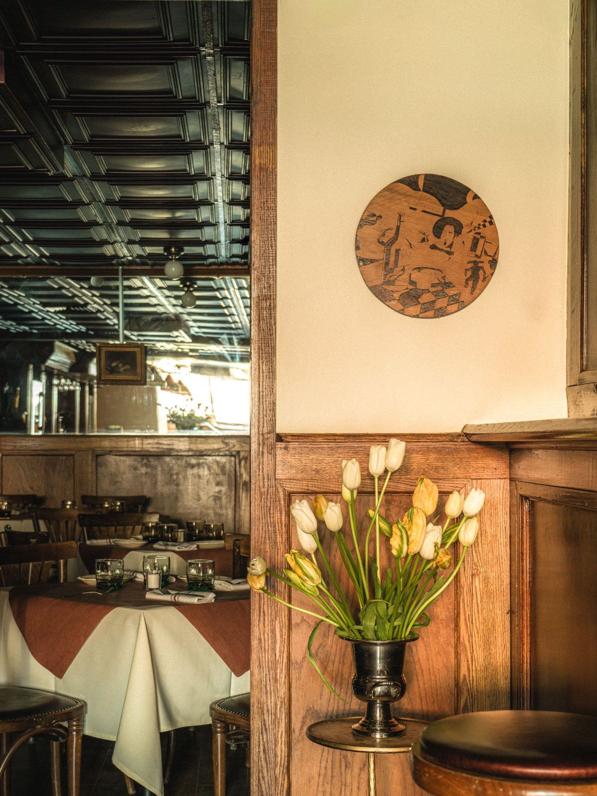 A cozy restaurant interior with tables set for dining, a tulip arrangement, and a decorative wooden wall art piece. Warm, inviting atmosphere.