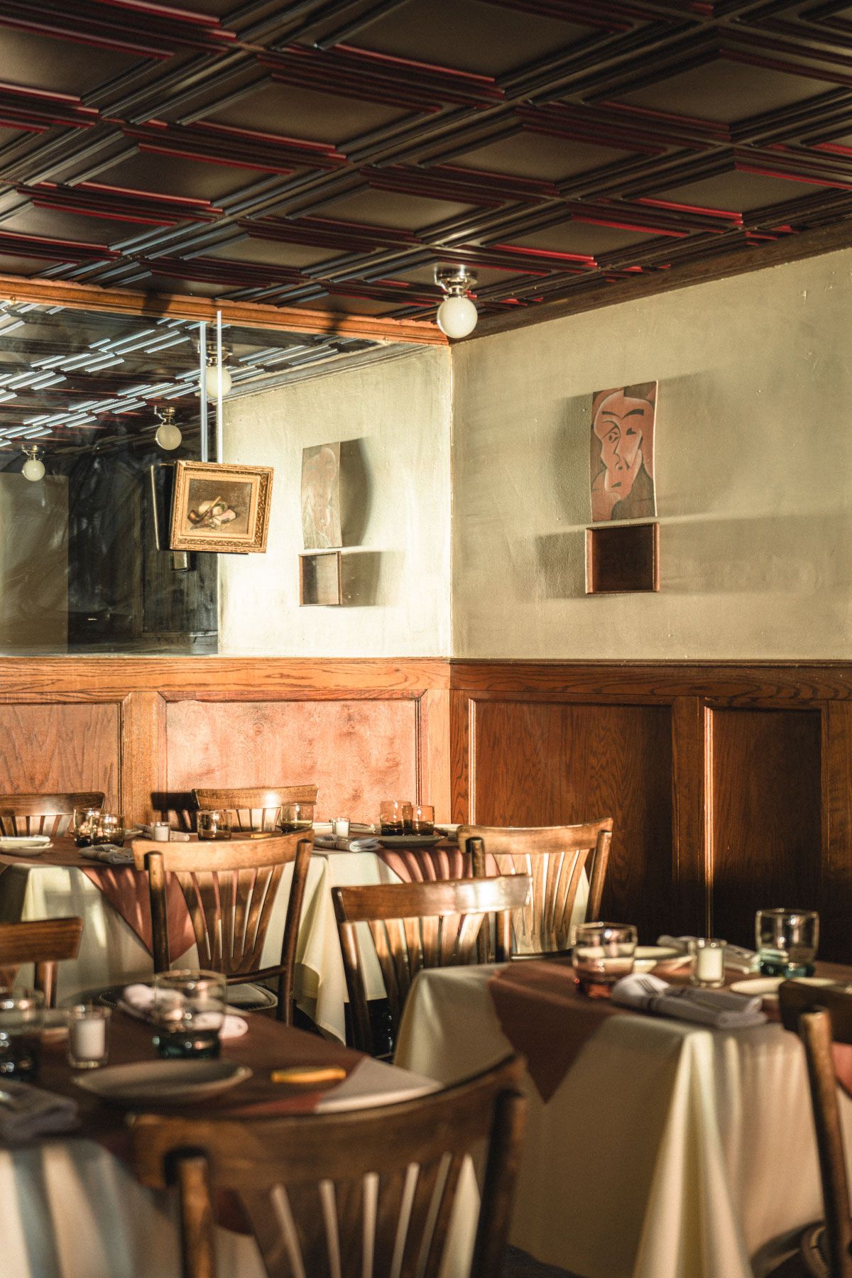 Cozy restaurant interior with wooden chairs, white tablecloths, and framed artwork on cream walls, warmly lit by soft lighting.