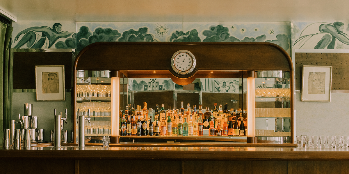 A vintage bar with various liquor bottles, a clock above, surrounded by art deco mural and framed artwork on the walls.