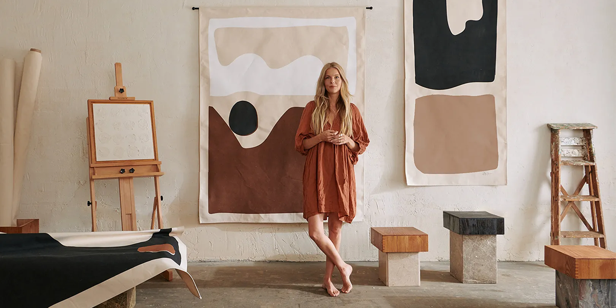 A woman stands in an art studio, surrounded by abstract paintings and minimalist wooden furniture, wearing a rust-colored dress.