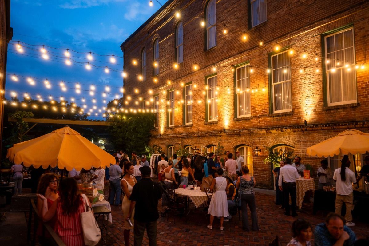 People socializing in a warmly lit courtyard party with string lights overhead, yellow umbrellas, and a brick building backdrop at dusk.