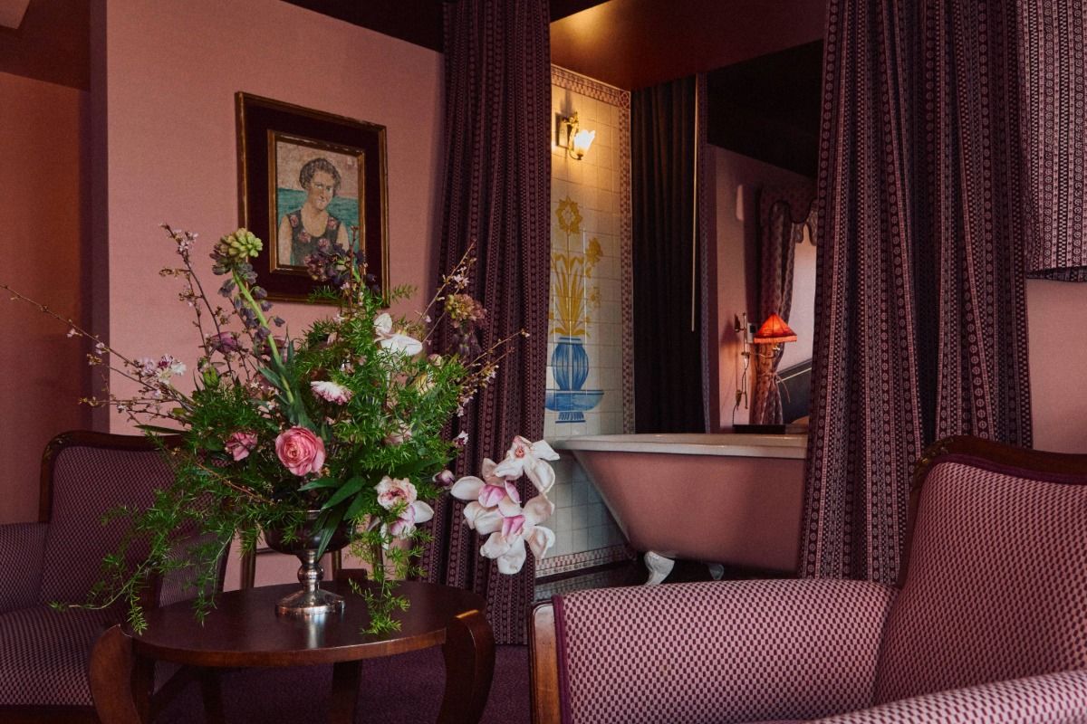 Cozy vintage-style room with pink walls, a floral arrangement, an elegant bathtub, patterned curtains, and a portrait on the wall.