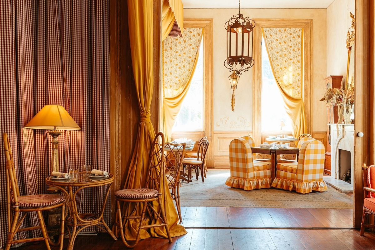 Cozy dining room with wicker furniture, checked upholstery, golden curtains, and vintage decor, illuminated by natural light and a table lamp.