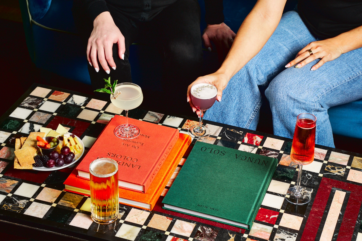 Two people seated with drinks on a colorful tiled table, alongside books and a platter of cheese, fruit, and crackers.