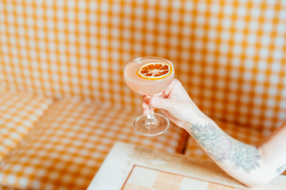 A tattooed hand holds a cocktail with a dried citrus slice garnish, set against a blurry orange gingham background.