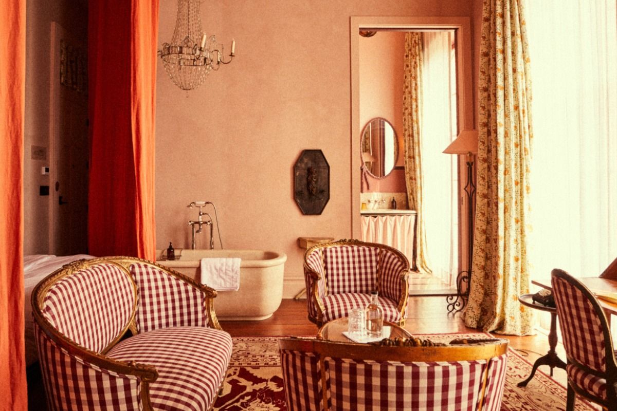 Elegant vintage-style room with red checkered seating, a freestanding bathtub, chandelier, and floral curtains by a sunlit window.