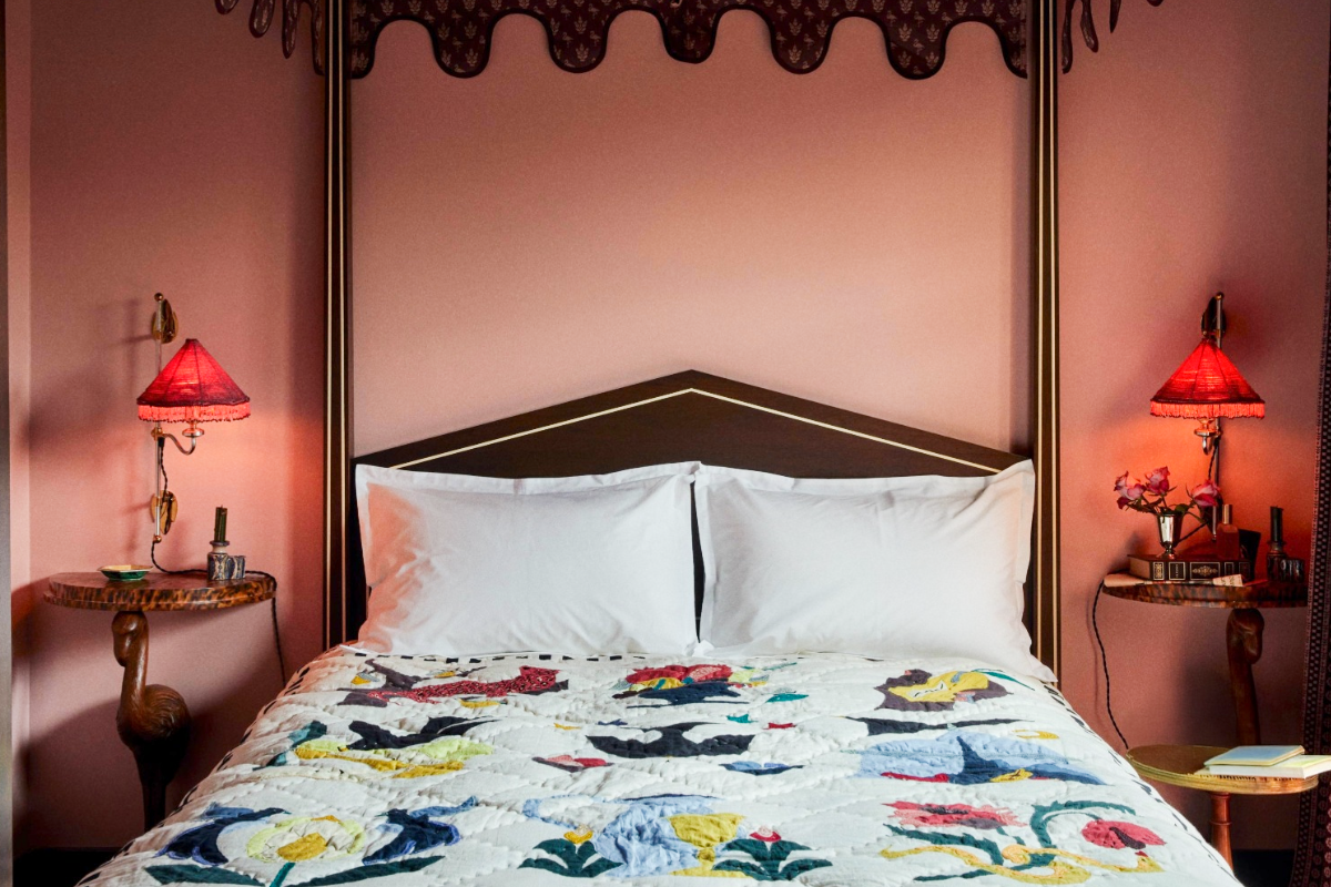 A cozy bedroom with a quilted bedspread, flanked by two round wooden tables with red lamps, set against a pink wall.